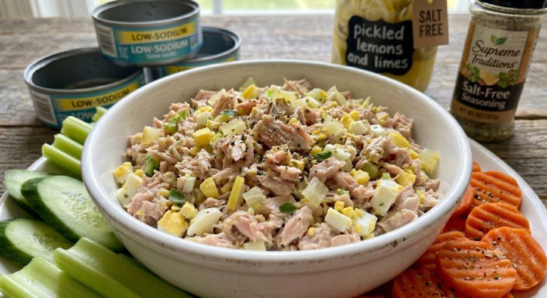 A clean white bowl filled with heart-healthy pickled lemon-lime tuna salad seasoned with marjoram, beautifully flanked by fresh cucumber slices, crisp celery sticks, and crinkle-cut carrot coins on a rustic wooden table landscape.